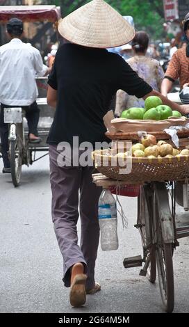 Eine Person, die einen Strohhut trägt, geht mit einem Fahrrad durch die Straßen von Hanoi, Vietnam, mit Körben mit Äpfeln auf der Rückseite des Fahrrads. Stockfoto