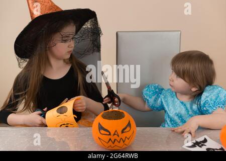 Zwei Mädchen Schwestern in Kostümen einer Hexe und Schneewittchen, die an einem Tisch sitzen, schneiden eine Papiergirlande zur Dekoration für den Halloween-Urlaub aus Stockfoto