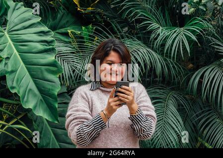 Porträt einer glücklichen Frau, die ihre sozialen Netzwerke auf ihrem Smartphone im Freien auf einem Hintergrund aus Laub betrachtet. Telefongesellschaft Konzept Stockfoto