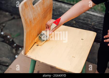 Makro-Kinderhand mit einem Pinsel malt einen kleinen alten Holzstuhl mit gelber Farbe Stockfoto