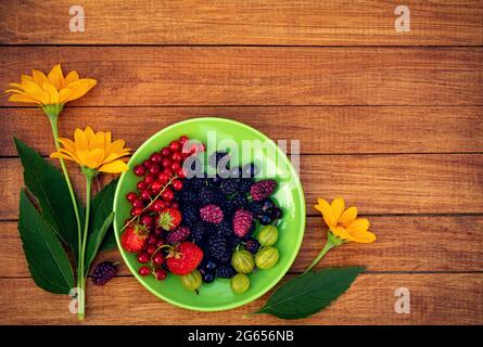 Berry mix - Johannisbeeren, Stachelbeeren, Maulbeeren, Erdbeeren auf einer grünen Untertasse mit gelben Blüten auf einem Holzhintergrund mit Platz für Design. Summe Stockfoto