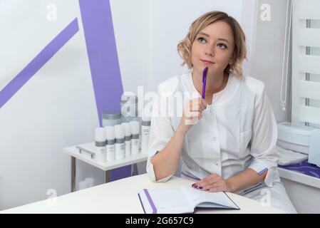 Arzt denkt lächelnd nach oben zu schauen. Kaukasische blonde Frau Arzt sitzt im Spa-Salon mit Bleistift und Notizbuch Stockfoto