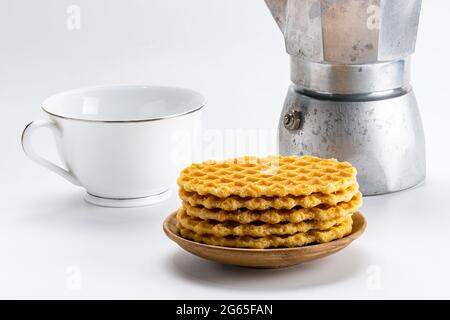 Stapel von knusprigen Waffeln auf einer Holzschale mit einer weißen Keramikschale und einer Mokka-Kaffeemaschine auf weißem Hintergrund mit Schneideweg. Stockfoto