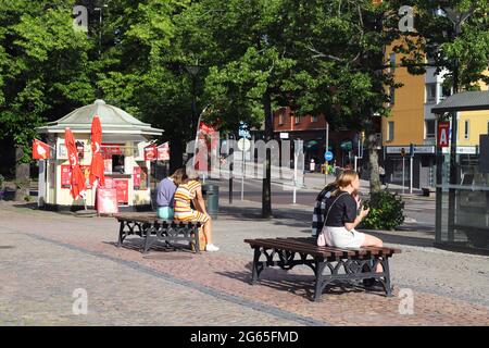 Karlstad, Schweden - 30. Juni 2021: Vier junge Menschen sitzen auf den Bänken auf dem Platz vor dem Eiskioské in der Sonne. Stockfoto