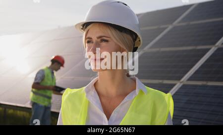 Eine schöne Solaringenieurin, die in der Nähe der Panels steht, und eine junge Spezialistin im Hintergrund testet die Batterien. Konzept des grünen Stroms. Stockfoto