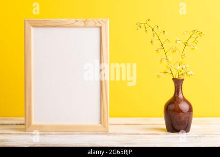 Holzrahmen mit gelben Barrenwort-Blüten in Keramikvase auf gelbem pastellfarbenem Hintergrund. Seitenansicht, Kopierraum, Stillleben, Mockup, Vorlage, Feder, Stockfoto