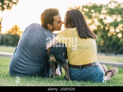 Rückansicht des jungen glücklichen Paares, das sich mit ihrem Hund im Park küsst Stockfoto