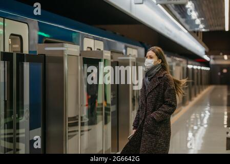 Eine Frau in einer medizinischen Gesichtsmaske, um die Ausbreitung des Coronavirus zu vermeiden, wartet auf einen ankommenden modernen Zug in der U-Bahn. Mädchen in einer chirurgischen Maske ist ke Stockfoto