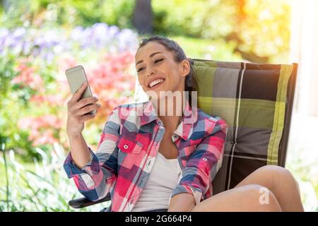 Attraktive Frau, die sich mit dem Smartphone im Stuhl zurücklehnt. Mädchen im Urlaub sitzen im Garten lächelnd entspannend. Stockfoto