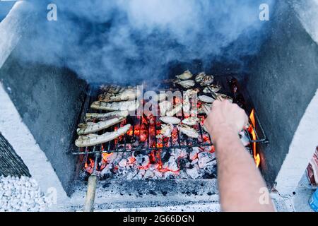 Detail der Hand eines Mannes mit einer Zange, die köstliches gegrilltes Fleisch über die Kohlen eines Grills dreht. Abendessen im Freien vorbereiten. Stockfoto