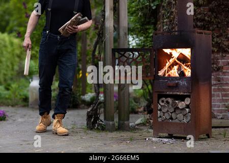 Ein Mann, der Holzstämme zu einem Metall brennenden Holzofen in seinem Garten trägt, während er das einfache Leben in einer ländlichen Gegend mit viel Grün lebt Stockfoto