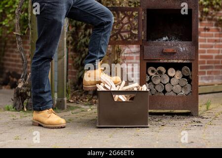 Mann in legerer Kleidung, der seinen Fuß auf einer Kiste voller gesammelter Holzstämme ruht, um als Brennstoff für seinen Gartenofen verwendet zu werden, während er sich entspannt Stockfoto