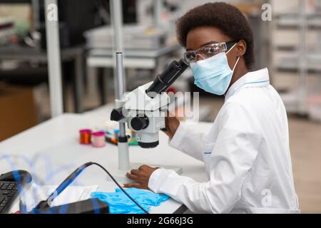 Wissenschaftler afroamerikanische Frau in Gesichtsmaske und Handschuhe arbeiten im Labor mit Mikroskop. Forschung und Entwicklung von medizinischen Chemikalien durch Farbe schwarze Frau. Stockfoto