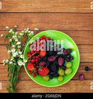 Beerenmischung - Johannisbeeren, Stachelbeeren, Maulbeeren, Erdbeeren auf einem Teller neben einem Strauß wilder Blumen auf einem hölzernen Hintergrund mit Kopierfläche. Sommer Stockfoto