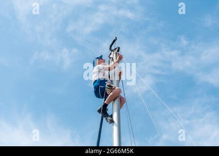 Mann in einem Sicherheitsgurt an der Spitze des Mastes einer Yacht oder eines Segelbootes, der Wartungsarbeiten an der Takelage durchführt, Großbritannien Stockfoto