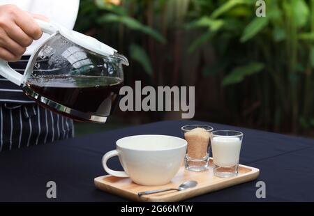Der Kellner gießt Kaffee in die Tasse. Kaffeetasse auf Holztablett mit Zucker und Milch daneben. Die Tischplatte ist weiß und liegt in der Gartenatmosphäre. Stockfoto