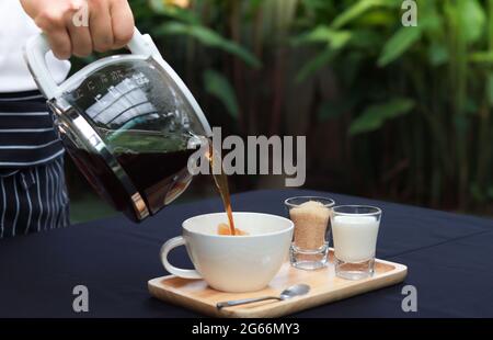 Der Kellner gießt Kaffee in die Tasse. Kaffeetasse auf Holztablett mit Zucker und Milch daneben. Die Tischplatte ist weiß und liegt in der Gartenatmosphäre. Stockfoto