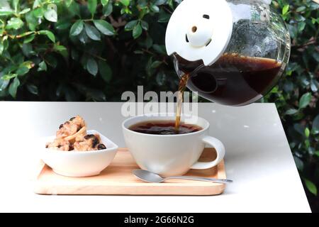 Gießen Sie den Kaffee aus der Kanne in die Tasse. Kaffeetasse auf Holztablett zusammen mit Keksen serviert. Tisch in Gartenatmosphäre mit grünen Blättern im Hintergrund. Stockfoto