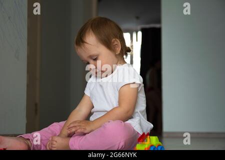 Kleines schönes einjähriges Mädchen in weißem T-Shirt und rosa Hosen wird mit Spielzeug im Zimmer gespielt. Stockfoto