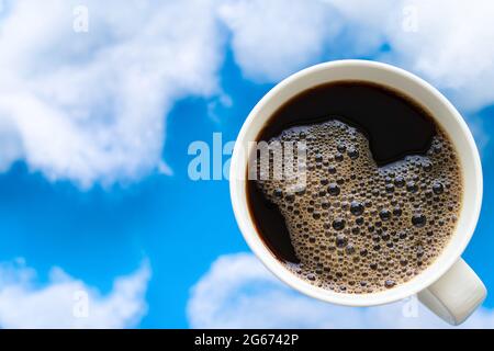High-Angle-Ansicht von schwarzem Kaffee mit Blase auf der Oberseite. Verschwommener blauer Himmel und weiße Wolken als Hintergrund. Speicherplatz kopieren. Stockfoto