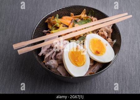 Schwarze Schüssel mit Ramen-Nudeln mit gekochten Eiern, Gemüse und truthahn treffen aufeinander, bereit, mit kochender Brühe gegossen zu werden. Essstäbchen aus Holz. Frisch zubereitet und Stockfoto