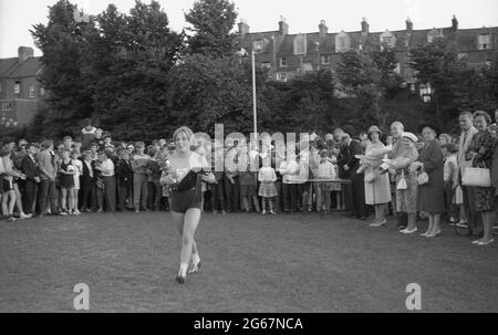 1960s, historisch, Leichtathletik, Schulsport zwischen den Bezirken, Preisverleihung, Eine siegreiche Sportlerin, die vor einer Gruppe anderer Athleten, Beamter und Eltern läuft, nachdem sie ihre Trophäe und Plakette erhalten hat, England, Großbritannien. Stockfoto