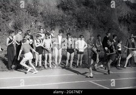 1960s, historischer Tag, Leichtathletik, Sporttag zwischen den Bezirken, Teenager-Jungen, die beim Staffellauf-Rennen gegeneinander antreten, den Staffelstab an den wartenden Läufer übergeben, beobachtet am Rande der Schlagerbahn von anderen Jungen, Devon, England, Großbritannien. Stockfoto