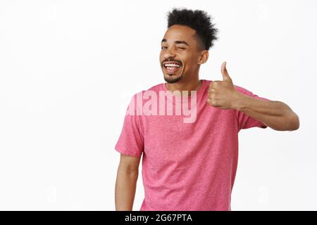 Gute Wahl. Lächelnder afroamerikanischer Kerl zwinkert einem zu, zeigt den Daumen vor Zustimmung, lobt großartige Arbeit, hervorragende Arbeit, steht in pinkem T-Shirt Stockfoto