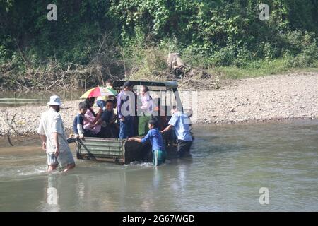 Männer schieben einen festgefahrenen Jeep durch einen Fluss mit einer Familie von Frauen und Kindern im Rücken, Luang Namtha, Laos Stockfoto