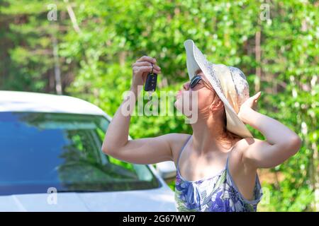 Schöne fröhliche Frau vor einem weißen Auto mit einem Schlüssel in der Hand an einem sonnigen Sommertag vor dem Hintergrund von grünen Bäumen. Selektiver Fokus Stockfoto