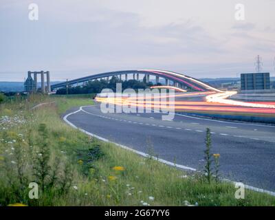 Elmley, Kent, Großbritannien. Juli 2021. Heute jährt sich die Eröffnung des Sheppey Crossing in Kent zum 15. Mal. Die Brücke wurde am 3. Juli 2006 als Teil eines £100m-Verbesserungsplans für die A249 zwischen der M2 und Sheerness Port (ein wichtiger Importeur von Autos) eröffnet. Kent Online berichtet, dass Highways England sagen, dass in den letzten 12 Monaten durchschnittlich 34,060 Fahrzeuge die Überfahrt täglich genutzt haben. Bild: In-Camera Composite Langzeitbelichtung zeigt Lichtspuren im Auto, die ältere Kingsferry Bridge ist links im Bild zu sehen. Kredit: James Bell/Alamy Live Nachrichten Stockfoto