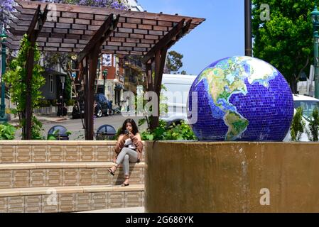 Junge Frau am Telefon am Wahrzeichen blauen Mosaikglobus Wasserbrunnen auf der Piazza Basilone in Little Italy, umgeben von Restaurants, Geschäften und Ferienwohnungen Stockfoto