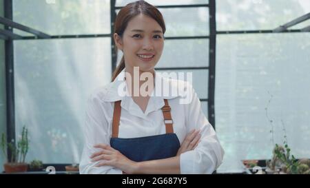 Asiatische schöne junge Gärtnerin Frau zuversichtlich mit Schürze stehend posiert gekreuzte Arme und lächelt vor der Kamera in Gewächshaus Kaktus Pflanzenkindergarten, klein Stockfoto
