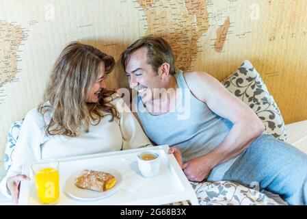 Reifes Paar lachte im Bett und hielt ein Tablett mit Frühstück. Stockfoto