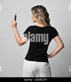 Blonde lockige Frau in schwarzem T-Shirt und weißer Hose steht mit dem Rücken zur Kamera und hält permanent Make-up-Maschine Stockfoto