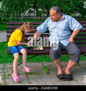 Kleines Mädchen spielt Schach mit Vater im Park auf einer Bank an der frischen Luft. Schachtag. Lehrreiche, lehrreiche Spiele im Freien. Entwicklung von Kindern. Stockfoto