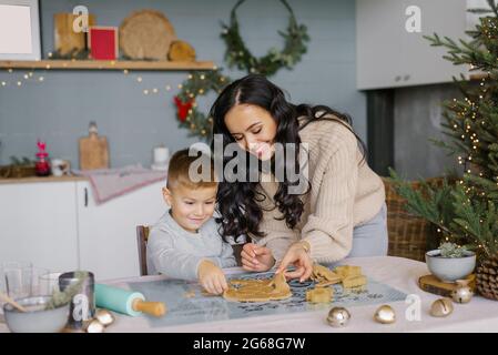 Eine fröhliche, schöne Mutter mit einem jungen Sohn schneidet in der Küche Weihnachtskekkkchen aus dem Teig und lacht. Stockfoto