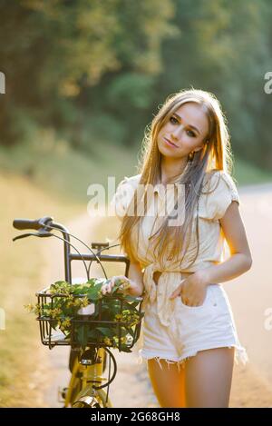 Junge schöne Frau, die neben einem Fahrrad mit einem Korbkorb voller Blumen im Wald steht Stockfoto