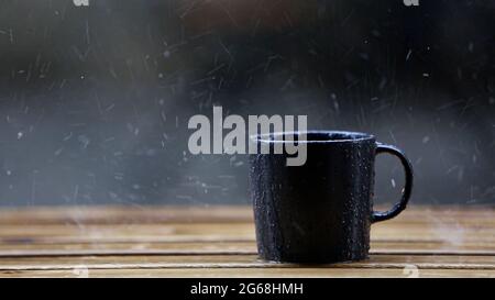 Schwarzer Kaffeebecher, der bei regnerischem Wetter auf Holzboden steht Stockfoto