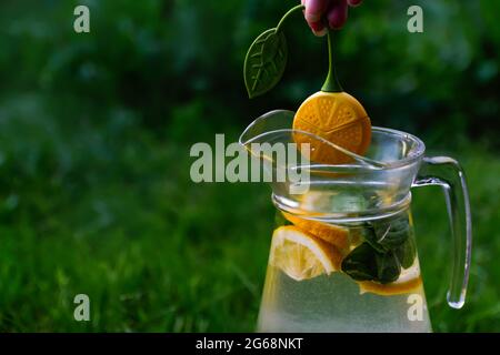 Defocus Hand hält Dummy Zitrone hängen über Glas Krug Limonade mit Scheibe Zitrone und Blätter der Minze auf natürlichem grünem Hintergrund. Krug des Sommers Stockfoto