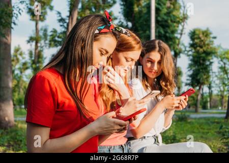 Drei Freundinnen beobachten Handys und haben Spaß auf der Bank im öffentlichen Park sitzen. Gen z, Jugendliche Geräte sucht, Digital Detox Stockfoto