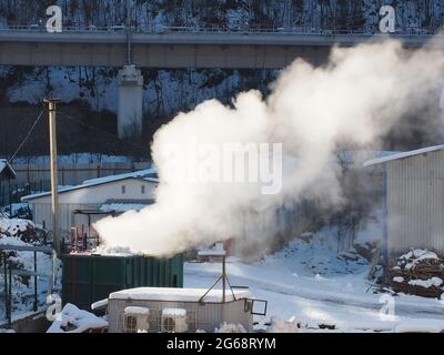 Im Winter auf einem Industriegelände vor dem Hintergrund einer Eisenbahnbrücke über einem grünen Eisencontainer dichter weißer Rauch Stockfoto