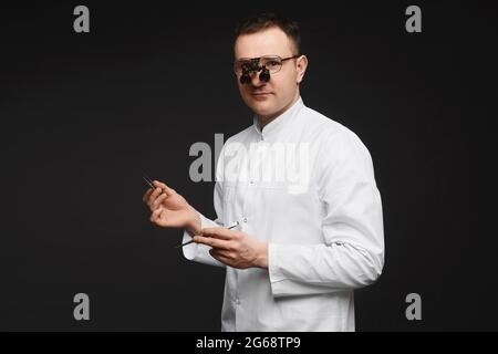 Kaukasischer Arzt in binokularen Lupengläsern und mit Chirurgen-Ausrüstung auf schwarzem Hintergrund. Junger Arzt Chirurg mit Ausrüstung für Stockfoto