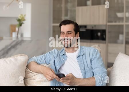 Überglücklich lachender junger Mann lenkte vom Smartphone ab, das auf der Couch saß Stockfoto
