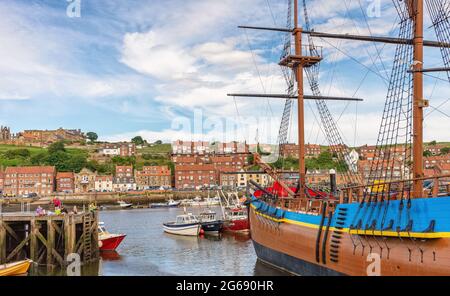 Eine Nachbildung der HMS Endeavour, die in Whitby Harbour vor Anker liegt. Eine Familie fischt von einem Steg aus und Gebäude säumen das Ufer. Ein wolkiger Himmel ist oben. Stockfoto