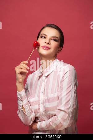Attraktive schöne junge Frau, die in der Hand rotes kleines Herz auf rotem Studioportrait hält Stockfoto