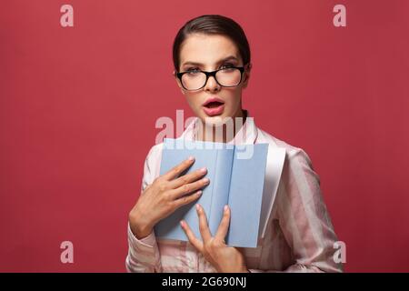 Schockiert Brünette Frau in Brille halten Buch posiert isoliert auf rotem Hintergrund Stockfoto