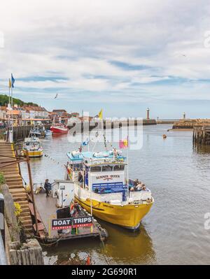 Stufen führen hinunter zu einem gelben Ausflugsboot, das in einem Hafen festgemacht ist. Zwei Leuchttürme und andere Boote sind in der Ferne und ein Himmel mit Wolken ist oben. Stockfoto