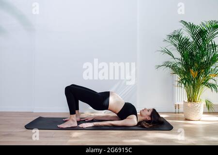 Blick von der Seite auf eine Schwangerin, die allein Yoga praktiziert und in einem großen Studio eine halbe Brücke macht. Sie trägt ein schwarzes Oberteil und eine Hose Stockfoto