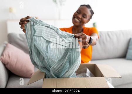 Zufriedener Käufer. Lächelnde schwarze Frau entpackt Paket nach der Lieferung, hält Bluse, sitzt auf der Couch zu Hause Stockfoto
